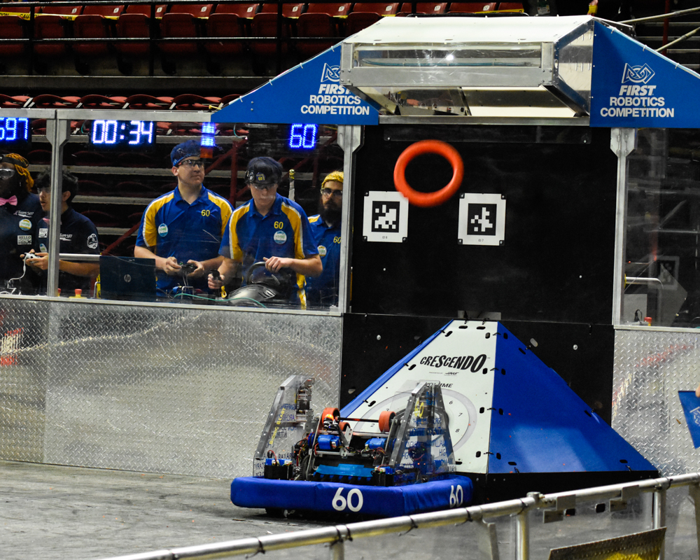 2024 robot scoring a game piece into the goal with the drive team visible through the glass next to the robot.